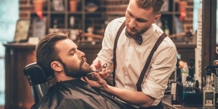 beard grooming side view of young bearded man getting beard haircut by hairdresser while sitting in chair at barbershop