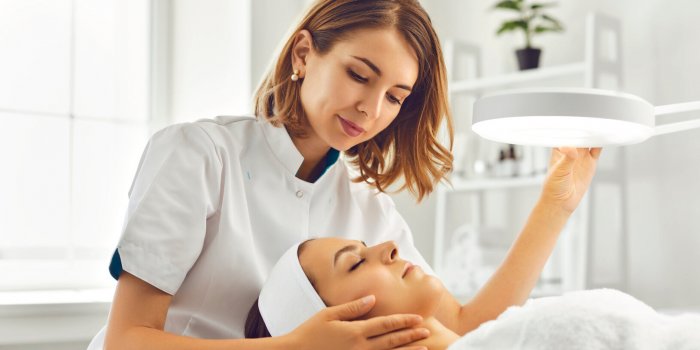 young woman cosmetologist or dermatologist directing lamp for facial treatment to lying young woman in beauty spa salon f...