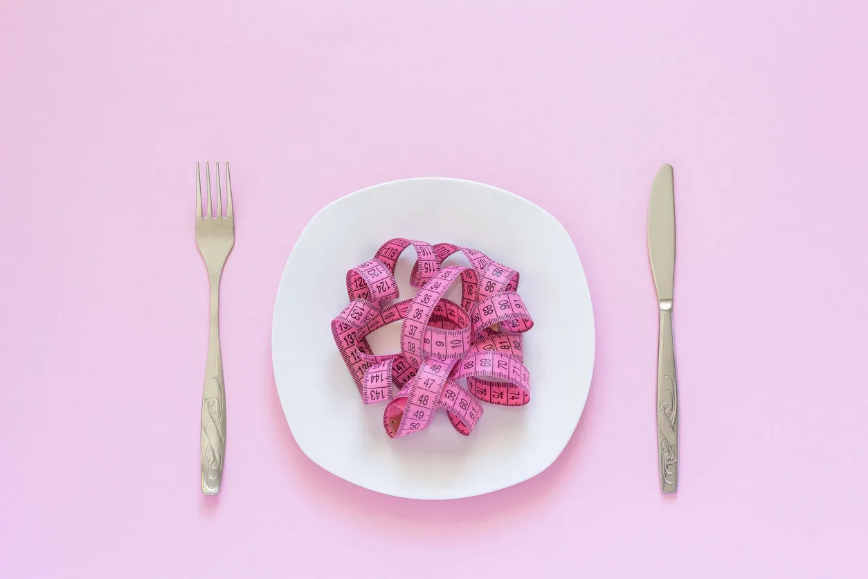 pink measuring tape lying on plate in the form of spaghetti, knife and fork on pink background diet or anorexia concept top view