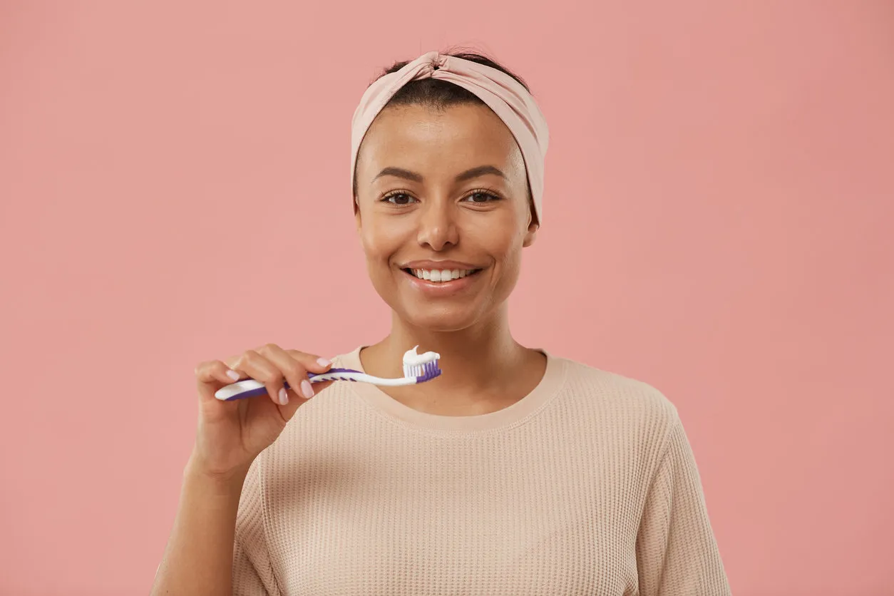 head and shoulders portrait of beautiful mixed race woman holding toothbrush and smiling at camera in morning while standing against dust pink background, copy space