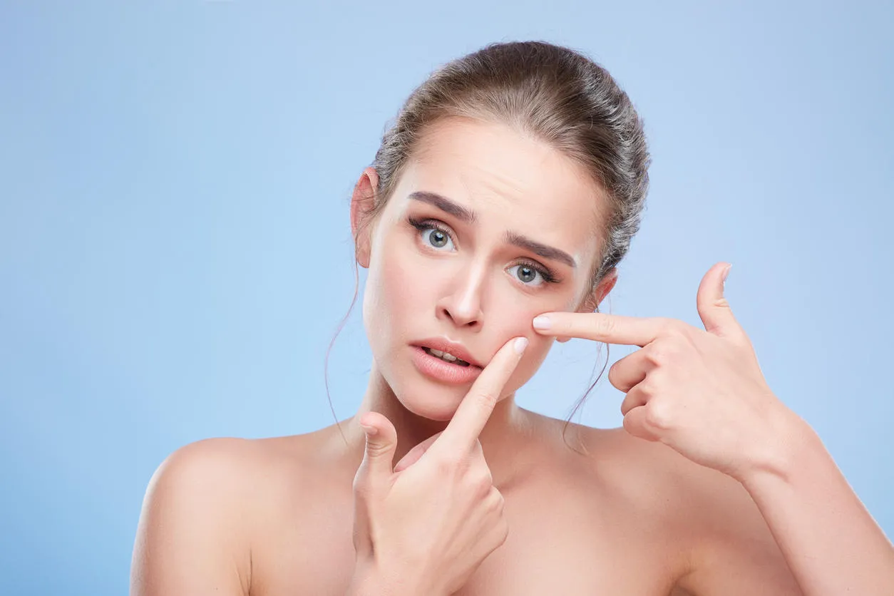beauty concept, head and shoulders of young woman with natural makeup disappointed girl looking at camera sadly and pressing out pimple on skin studio, indoors