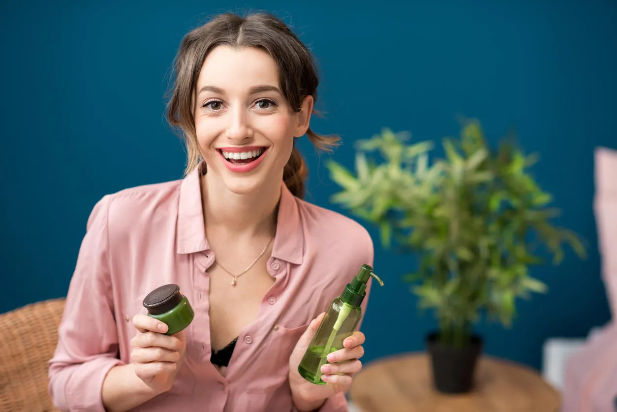 portrait of a young woman holding green bottles with lotion on the blue wall background at home
