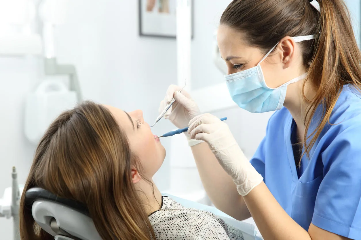 dentist wearing mask gloves and blue uniform examining a patient teeth with a dental probe and a mirror in a box with medical equipment in the background dentist wearing mask gloves and blue uniform examining a patient teeth with a dental probe and a mirror in a box with medical equipment in the background