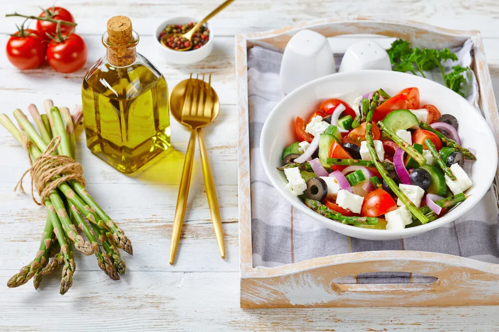 healthy grilled asparagus salad with feta cheese, tomatoes, cucumbers, black olives, red onion rings with olive oil served on a white bowl on wooden background on a serving tray with ingredients