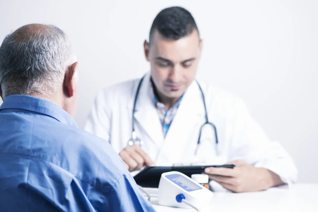 closeup of a senior caucasian patient man seen from behind and a caucasian doctor man checking his medical history or the results of a test in his tablet, sitting both at the doctors desk