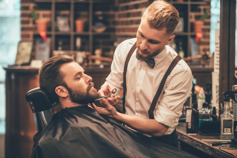 beard grooming side view of young bearded man getting beard haircut by hairdresser while sitting in chair at barbershop beard grooming side view of young bearded man getting beard haircut by hairdresser while sitting in chair at barbershop