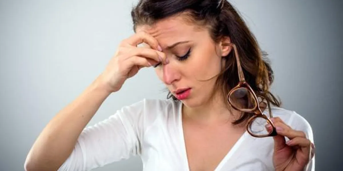 séduisante jeune femme avec un mal de tête tenant ses lunettes dans une main comme elle pince le pont de son nez avec une expression peinée, la tête et les épaules sur gris séduisante jeune femme avec un mal de tête tenant ses lunettes dans une main comme elle pince le pont de son nez avec une expression peinée, la tête et les épaules sur gris