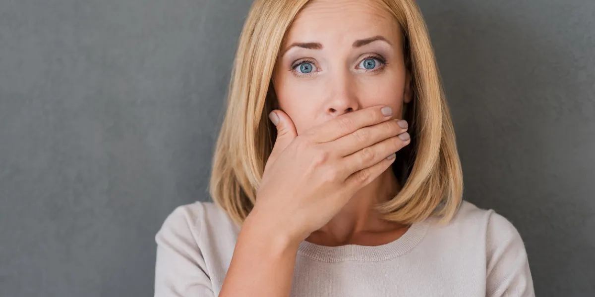 surprised mature woman covering mouth with hand and staring at camera while standing against grey background surprised mature woman covering mouth with hand and staring at camera while standing against grey background