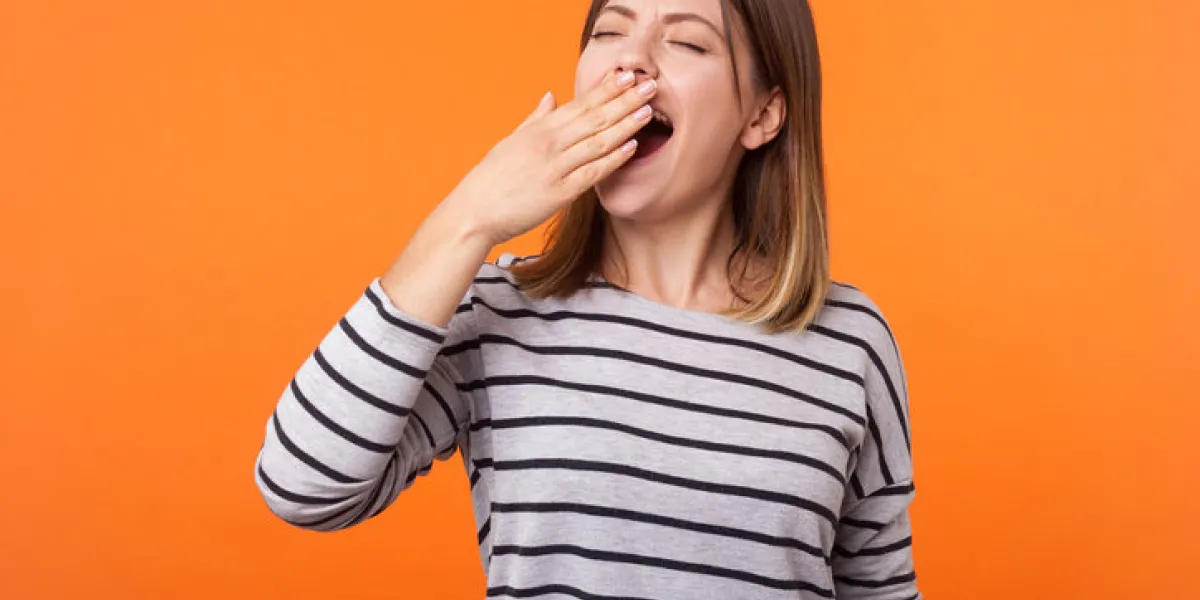 portrait of exhausted or bored young woman with brown hair in long sleeve striped shirt standing, yawning and covering mouth with hand, disinterest indoor studio shot isolated on orange background portrait of exhausted or bored young woman with brown hair in long sleeve striped shirt standing, yawning and covering mouth with hand, disinterest indoor studio shot isolated on orange background