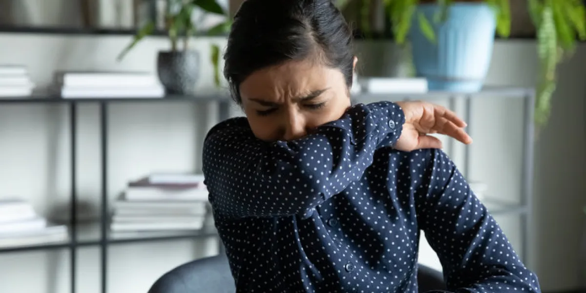 millennial indian ethnic girl sitting in office, coughing in elbow, right illness behavior for not spreading virus infection unhealthy young woman feeling unwell at workplace, covid 19 symptoms