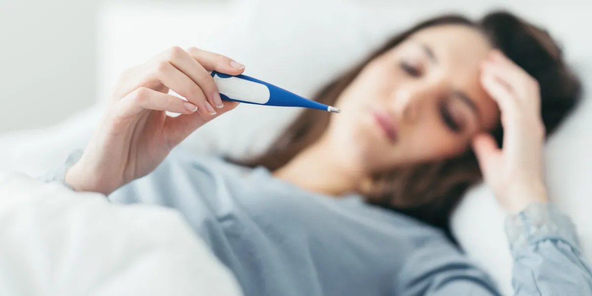 woman with flu virus lying in bed, she is measuring her temperature with a thermometer and touching her forehead woman with flu virus lying in bed, she is measuring her temperature with a thermometer and touching her forehead