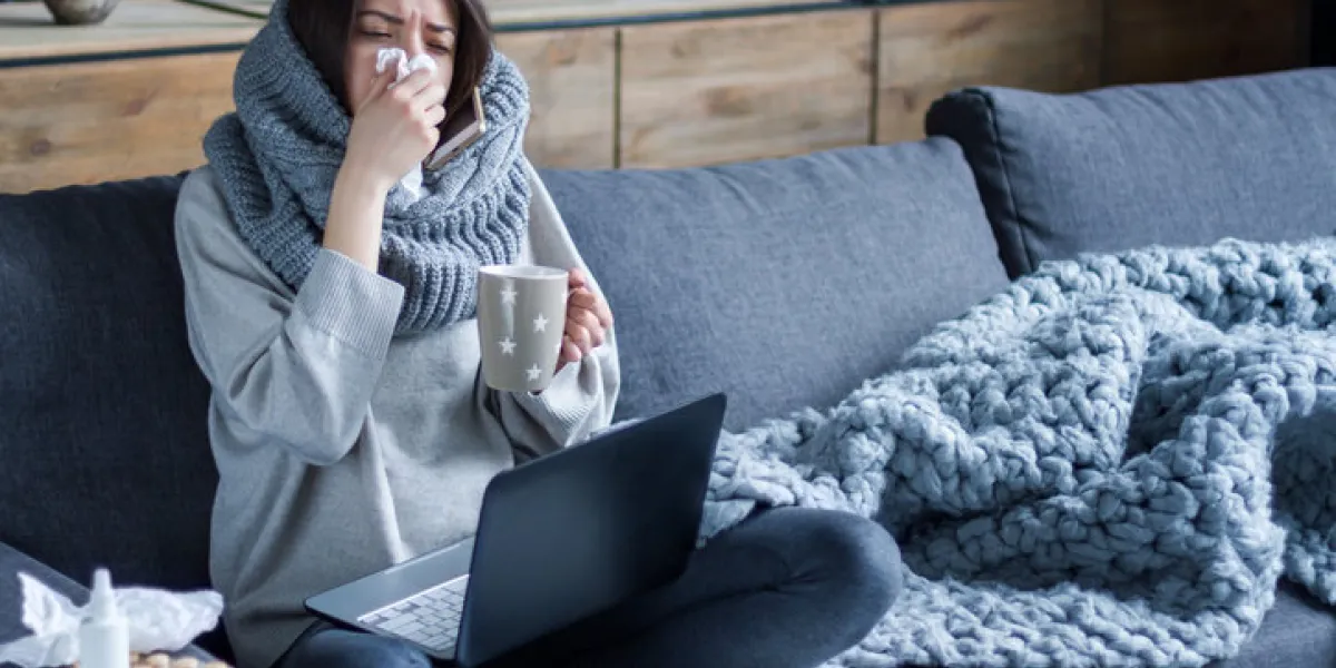 ill brunette girl is sitting at sofa and working remotely on laptop female is blowing out snot, having fever and headache young woman is treated at home, took sick leave winter cold and flu concept