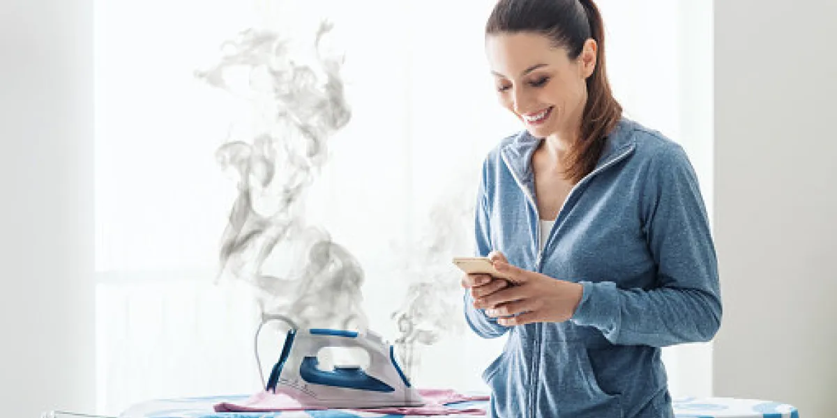 careless smiling housewife at home texting with her smartphone, she has left the iron on the ironing board careless smiling housewife at home texting with her smartphone, she has left the iron on the ironing board