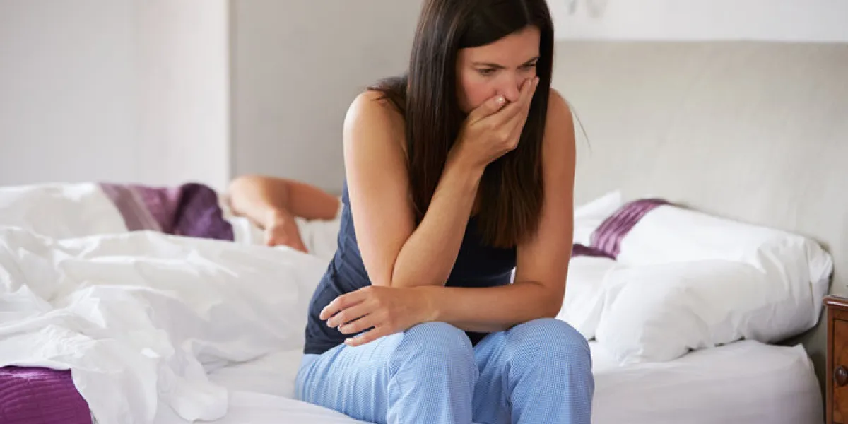woman sitting on bed feeling sick, while man sleeps woman sitting on bed feeling sick, while man sleeps