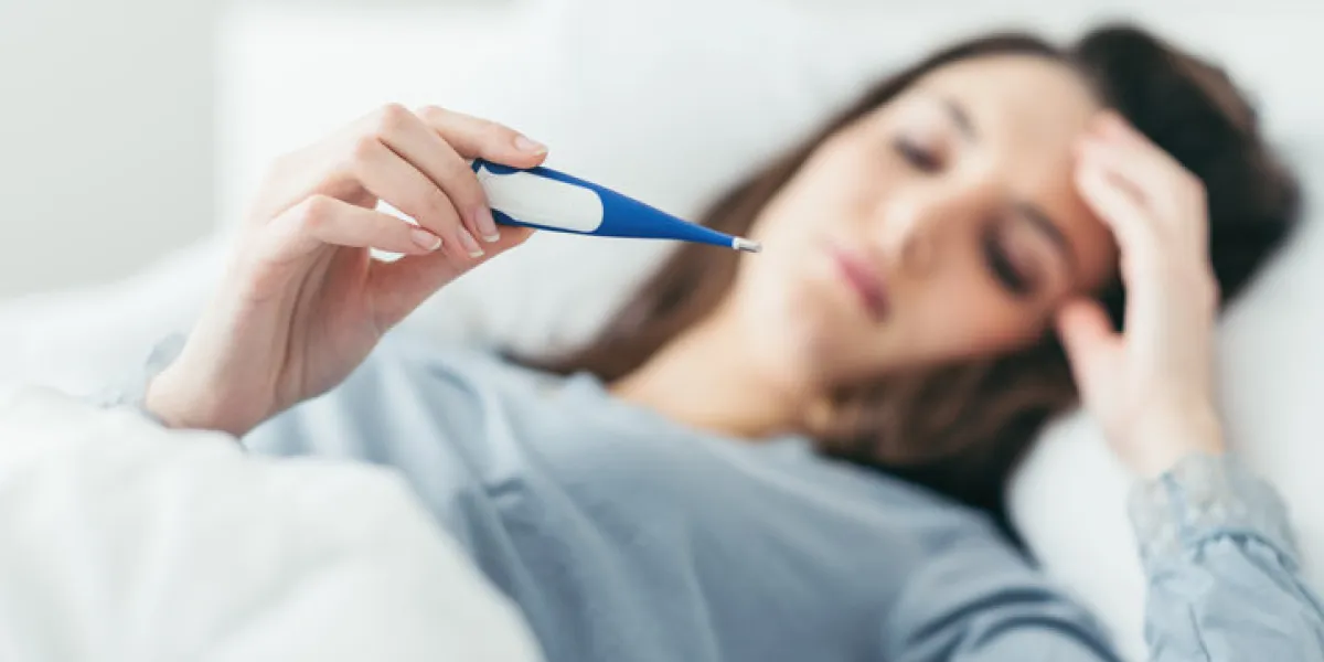 woman with flu virus lying in bed, she is measuring her temperature with a thermometer and touching her forehead woman with flu virus lying in bed, she is measuring her temperature with a thermometer and touching her forehead