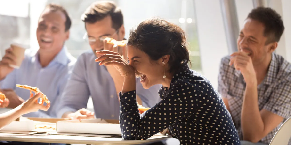 indian woman laughing at funny joke eating pizza with diverse coworkers in office, friendly work team enjoying positive emotions and lunch together, happy colleagues staff group having fun at break