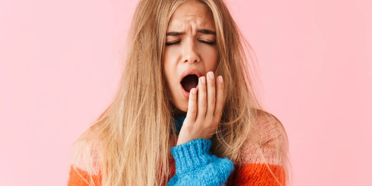 portrait of a tired young girl wearing sweater standing isolated over pink background, yawning