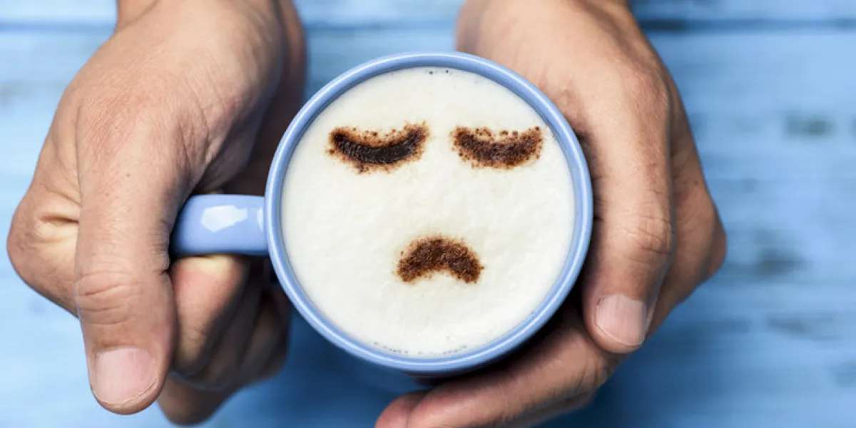 high-angle shot of a young caucasian man with a blue cup of cappuccino with a sad face drawn with cocoa powder on the milk foam, on a blue rustic table high-angle shot of a young caucasian man with a blue cup of cappuccino with a sad face drawn with cocoa powder on the milk foam, on a blue rustic table