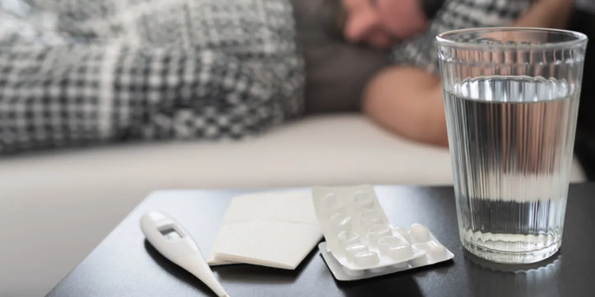 meds, medical thermometer and glass of water on nightstand against sick person suffering from flu or common cold sleeping in bed