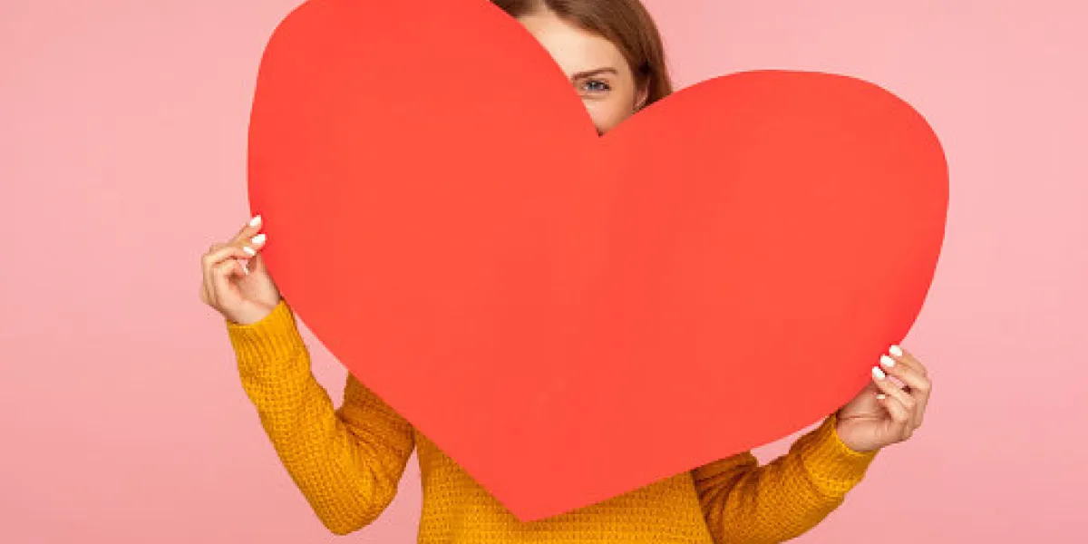 valentine's day portrait of funny girl hiding behind big red heart and looking at camera with playful eyes, holding symbol of love and affection indoor studio shot isolated on pink background