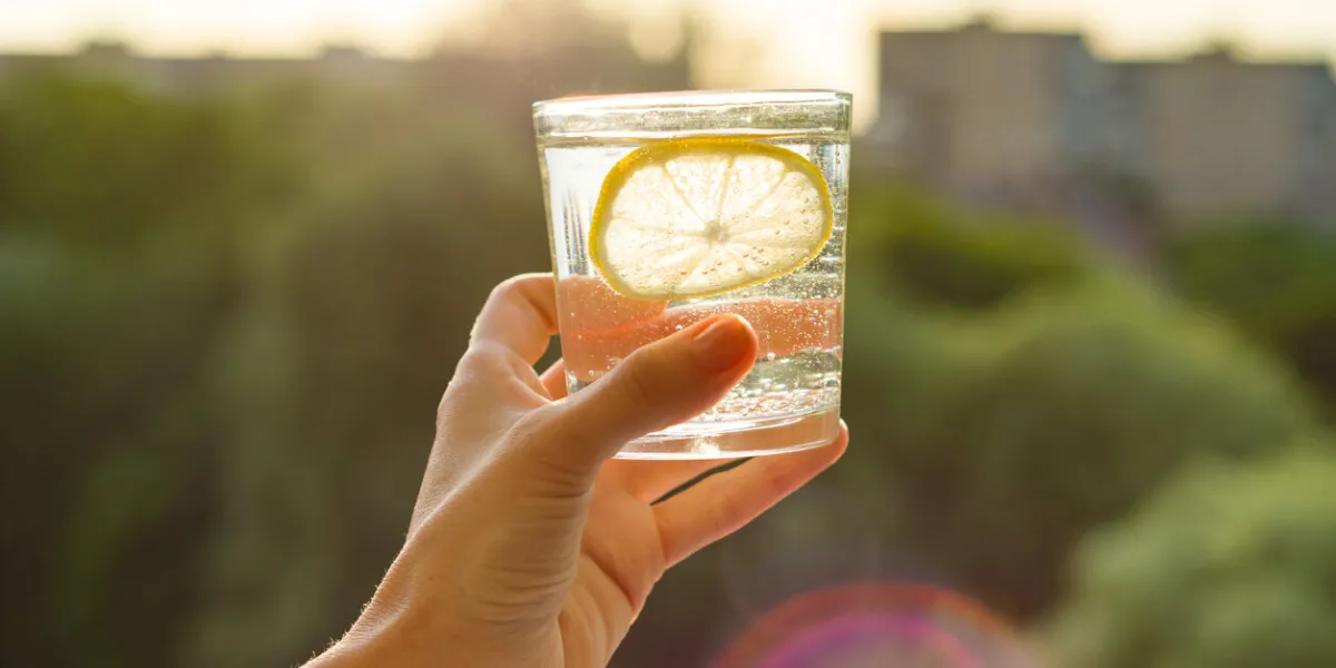 glass of clear, sparkling water with lemon in hand background sky, silhouette of the city, sunset