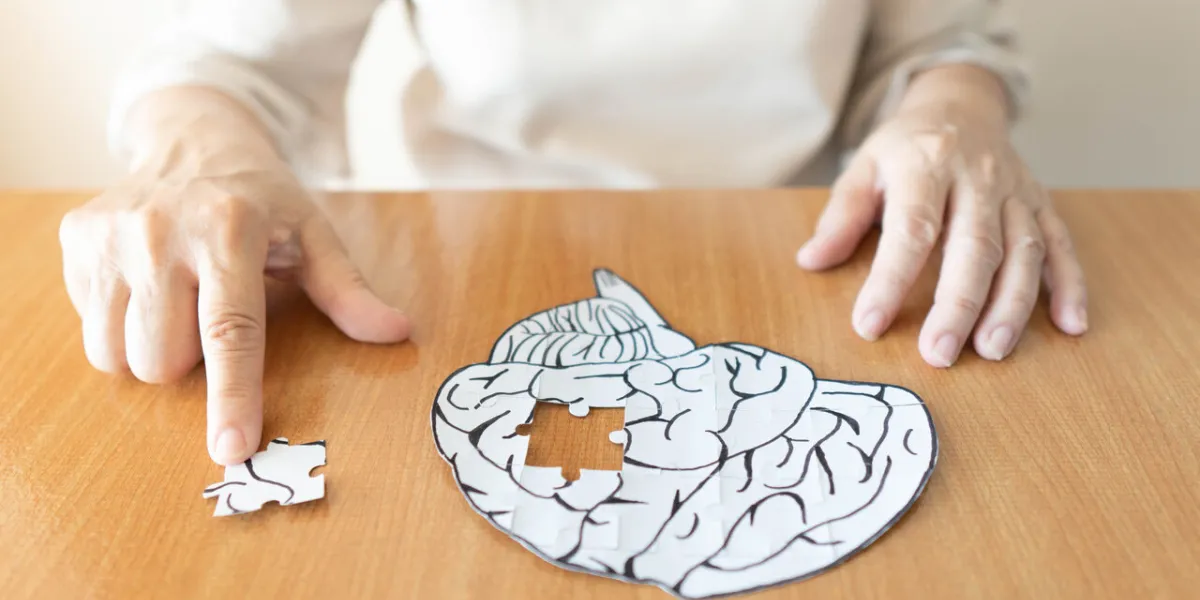 elderly woman hands putting missing white jigsaw puzzle piece down into the place as a human brain shape creative idea for memory loss, dementia, alzheimer's disease and mental health concept
