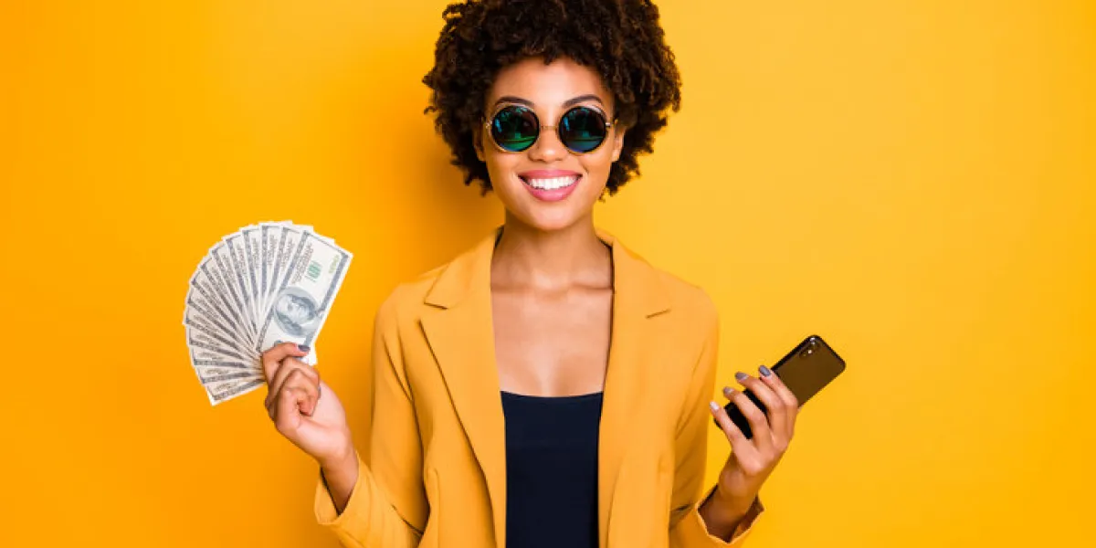 portrait of positive cheerful afro american lady freelancer get cashback, salary hold money fan use smartphone buy purchase pay online wear style blazer suit isolated over yellow color background