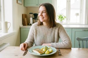 photo réaliste d'une jeune femme souriante, assise à la table de sa cuisine lumineuse, déjeunant ave