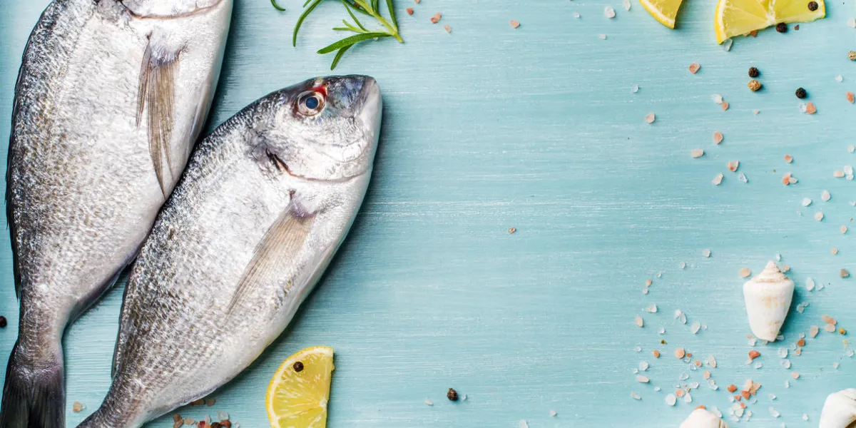 fresh raw sea bream fish decorated with lemon slices, herbs and sea shells on blue wooden background healthy food concept, top view, copy space