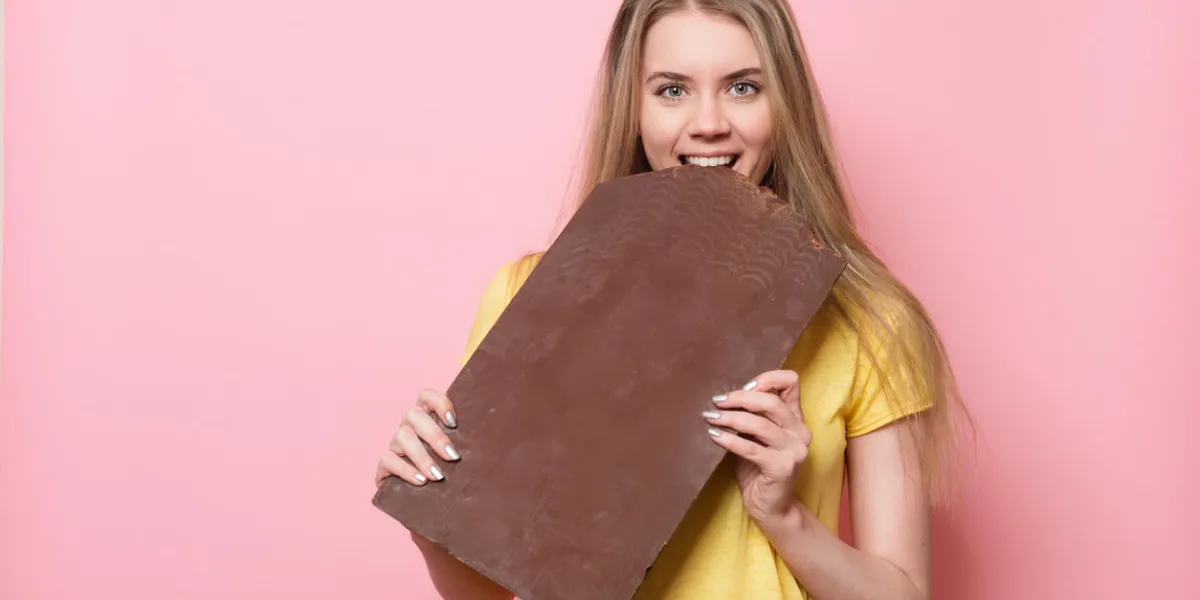 woman with chocolate smiling cute girl holding and eating giant cocoa chocolate bar near pink wall