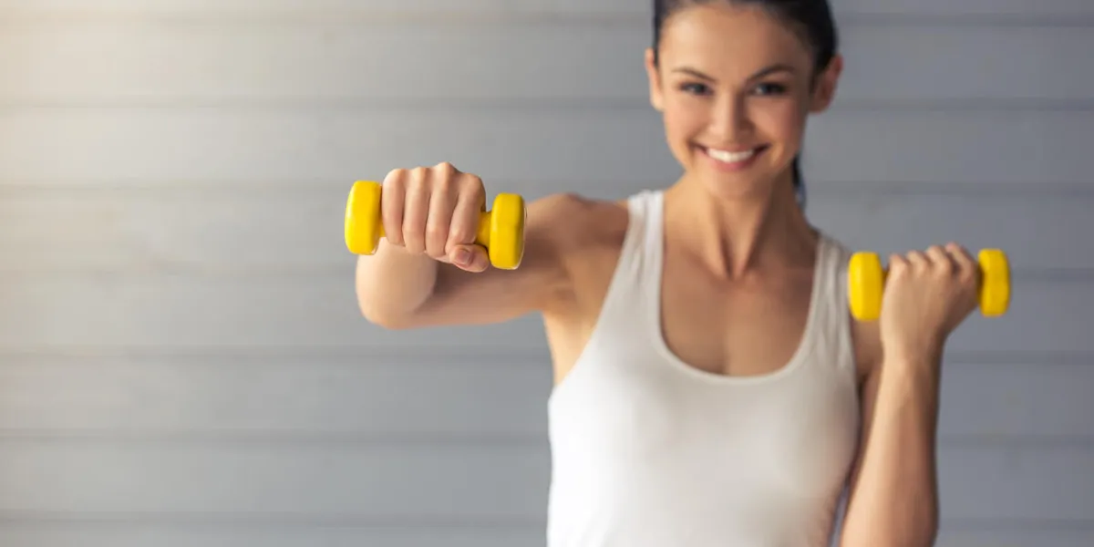 beautiful young woman in sports wear is working out with dumbbells, looking at camera and smiling, standing on a gray background beautiful young woman in sports wear is working out with dumbbells, looking at camera and smiling, standing on a gray background