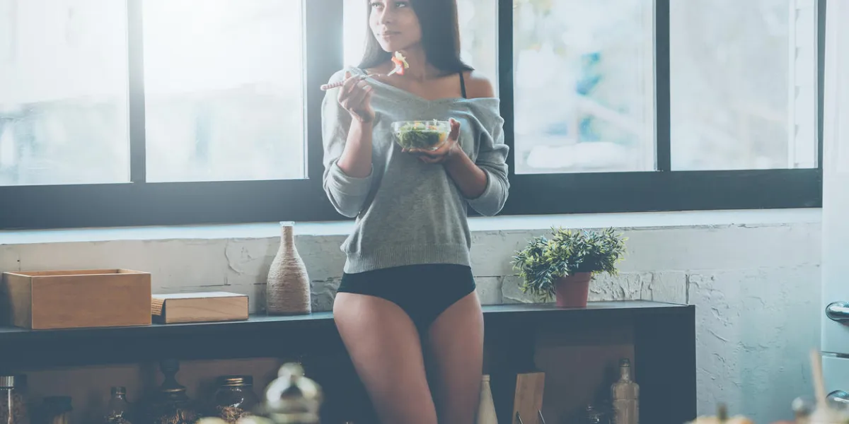belle jeune femme métisse, manger salade et regarder loin loin debout dans la cuisine à la maison belle jeune femme métisse, manger salade et regarder loin loin debout dans la cuisine à la maison