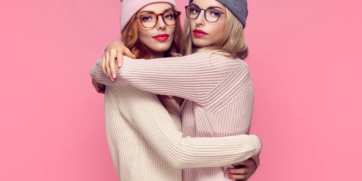 fashion young woman hugging in stylish autumn outfit pretty sisters best friends twins hipster blond redhead model, fashion cozy jumper, glasses beautiful girls in trendy beanie hat on pink