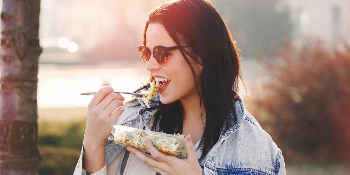 young brunette woman in sunglasses eating fresh salad outdoor in sunset, looking away young brunette woman in sunglasses eating fresh salad outdoor in sunset, looking away