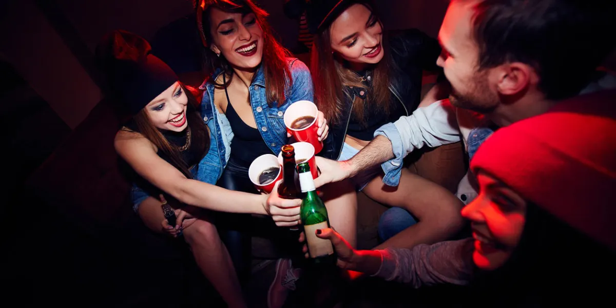 grand angle de plusieurs jeunes gens, filles et un homme, se saouler à la fête de club, assis près ensemble dans la pièce allumée rouge foncé et acclamant à leur réunion