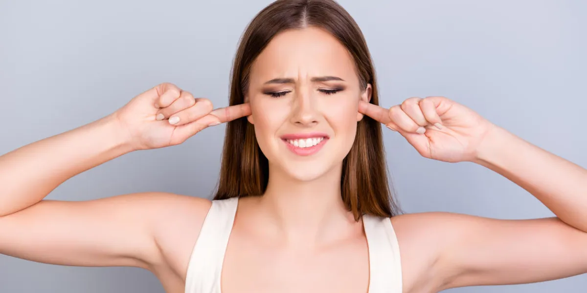 portrait of tired young cute girl, closing her ears with fingers she is with closed eyes also, in a white casual singlet on pure background