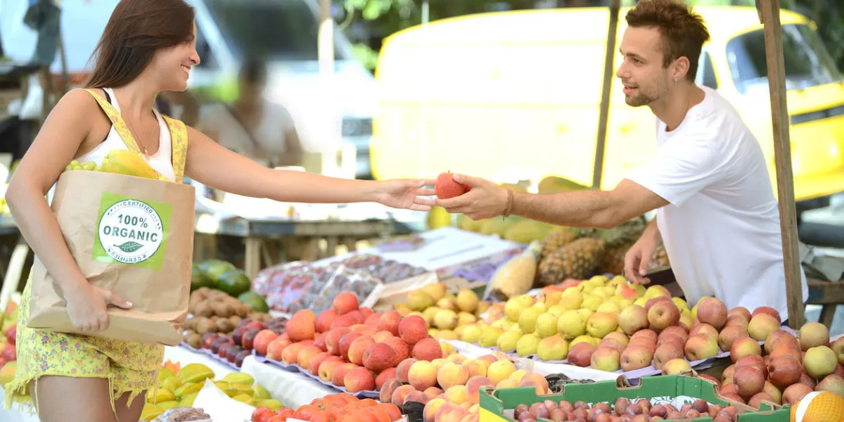 marchand de fruits et légumes, propriétaire d'une petite entreprise dans un marché ouvert, distribuant un fruit à un consommateur, portant un sac de papier commercial avec une étiquette certifiée biologique 100 pleine de fruits et légumes