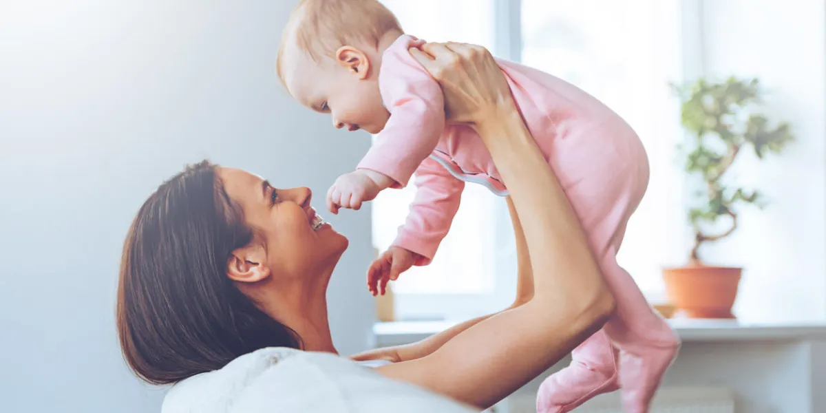 side view of cheerful beautiful young woman holding baby girl in her hands and looking at her with love while sitting on the couch at home side view of cheerful beautiful young woman holding baby girl in her hands and looking at her with love while sitting on the couch at home