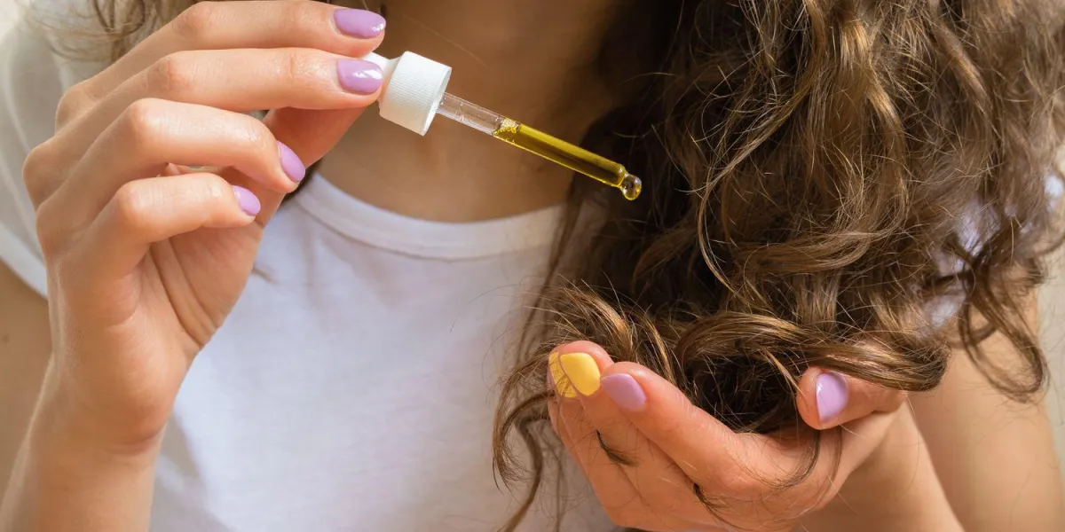 jeune fille en t-shirt blanc tient dans sa main gauche les bouts de ses cheveux bouclés et sa main droite leur applique l'huile jaune