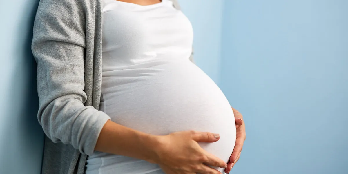 mid-section portrait of unrecognizable woman during last months of pregnancy holding her big belly gently standing against wall in blue room