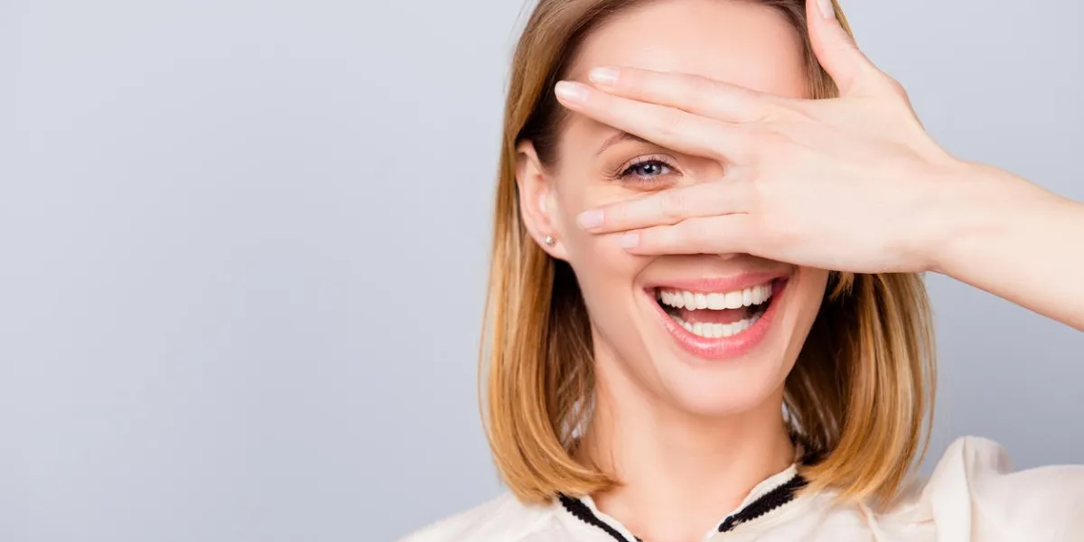 blond young woman with beaming smile is looking through her hand she is wearing casual outfit and stands on light grey background blond young woman with beaming smile is looking through her hand she is wearing casual outfit and stands on light grey background