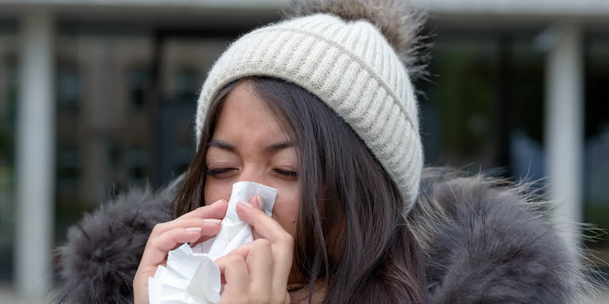 young woman with a seasonal winter cold or flu wearing a furry jacket and knitted cap blowing her nose on a white handkerchief outdoors on an urban street