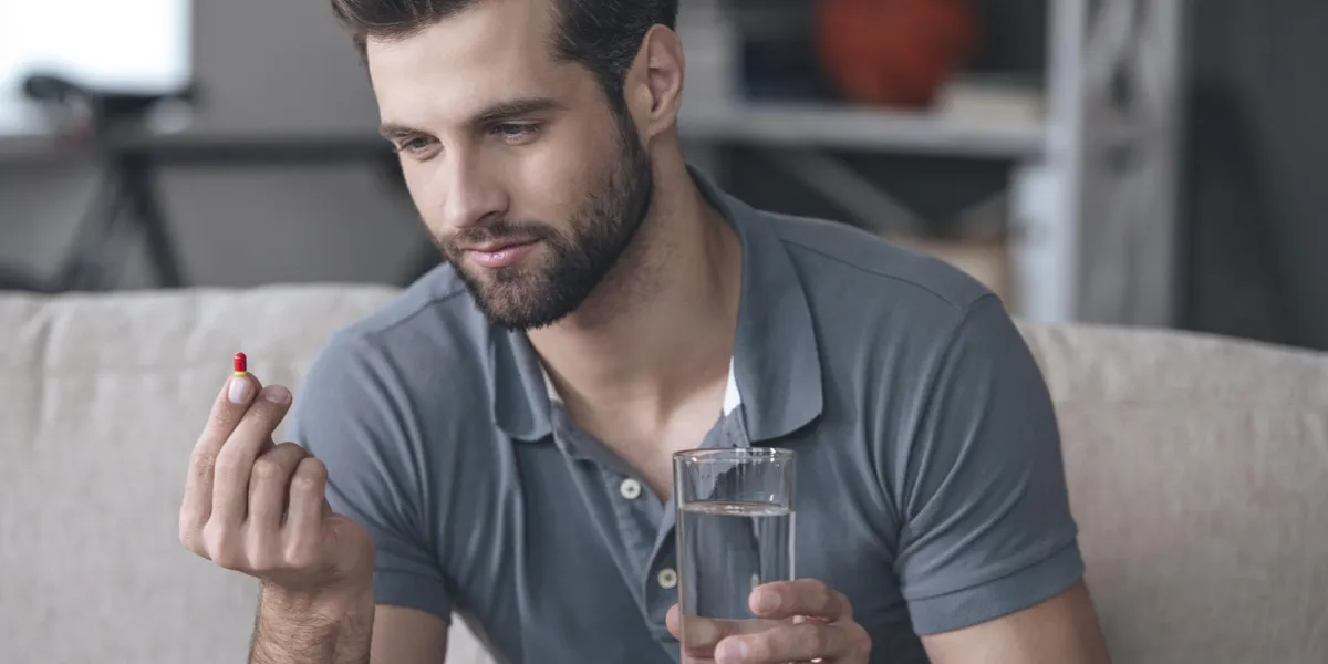 beau jeune homme tenant un verre d'eau et en regardant une pilule dans sa main tout en étant assis sur le canapé à la maison beau jeune homme tenant un verre d'eau et en regardant une pilule dans sa main tout en étant assis sur le canapé à la maison