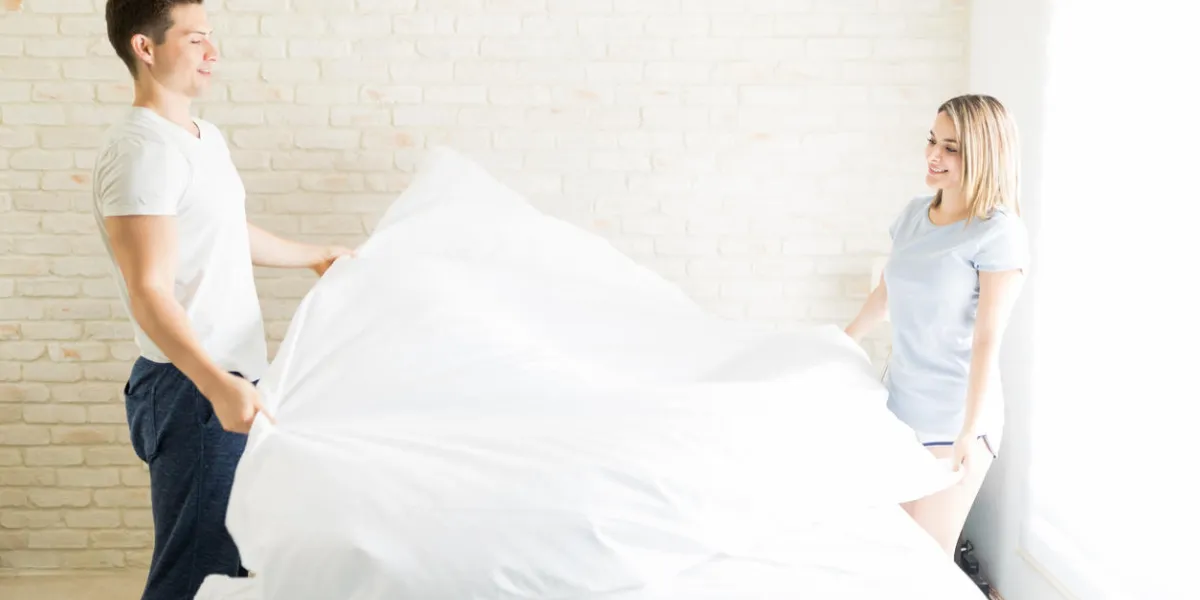 multiethnic lovers arranging white sheet on bed in apartment multiethnic lovers arranging white sheet on bed in apartment