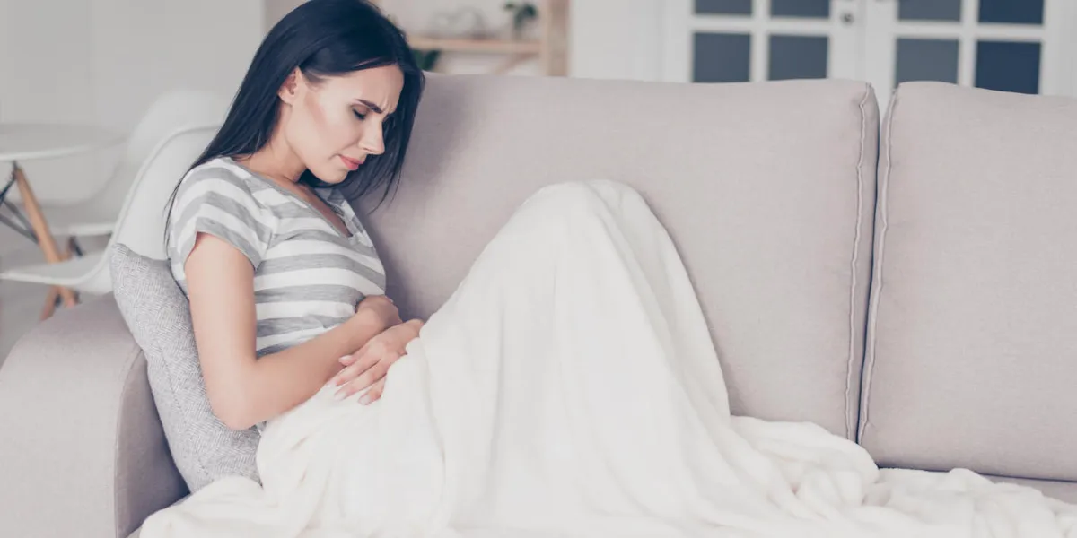sad beautiful woman sitting couch and feel spasm and symptoms of pms while touch belly with coverlet