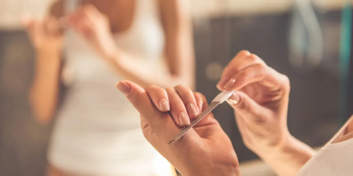 beautiful young woman in white undershirt is filing her nails and smiling while standing in front of the mirror in bathroom, close-up
