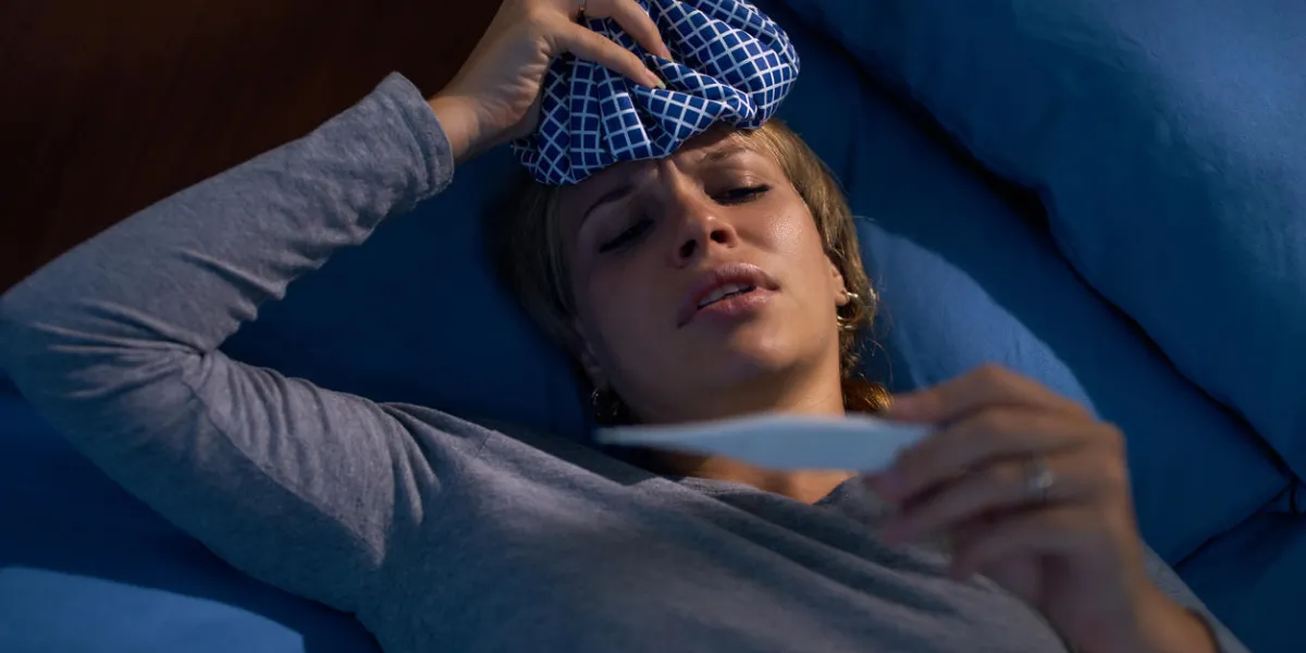 young hispanic woman in pajamas, laying on bed with fever and ice bag on head, measuring temperature with digital thermometer at night high angle view young hispanic woman in pajamas, laying on bed with fever and ice bag on head, measuring temperature with digital thermometer at night high angle view