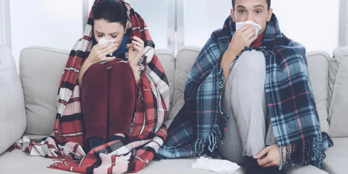 the couple is sitting on the couch wrapped in blankets man and woman are sick the man flies, the woman also blows her nose into a napkin