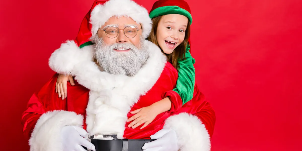 close up photo of charming santa claus in eyeglasses eyewear and his little elf helper in green hat headwear hugging piggyback isolated over red background close up photo of charming santa claus in eyeglasses eyewear and his little elf helper in green hat headwear hugging piggyback isolated over red background