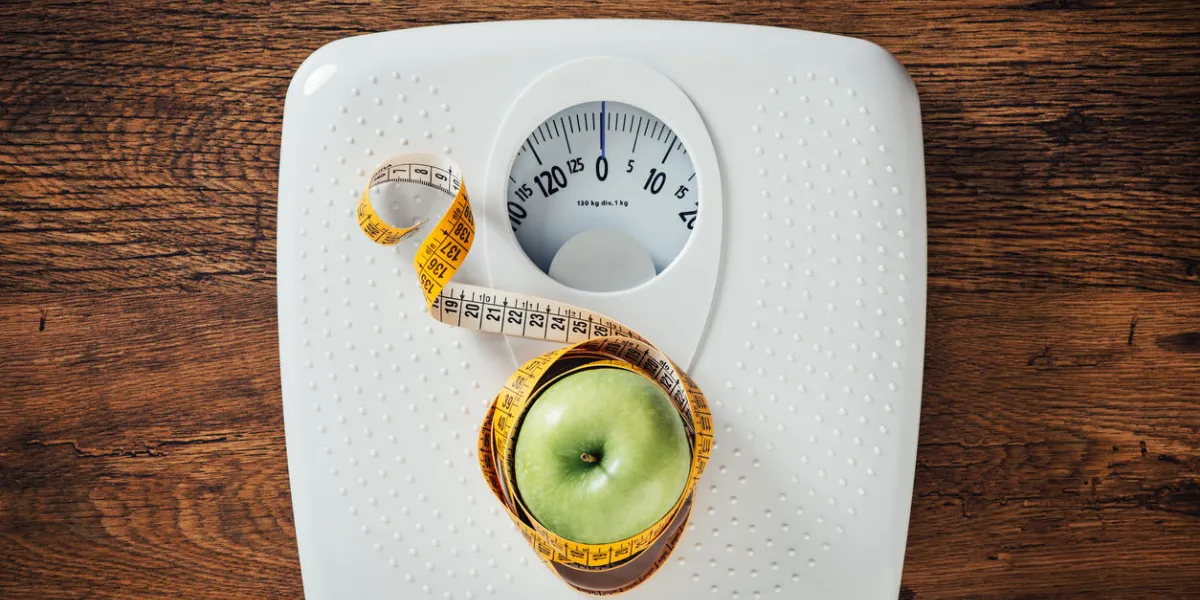 green apple wrapped in a tape measure on a white scale, wooden surface on background, dieting and weight loss concept